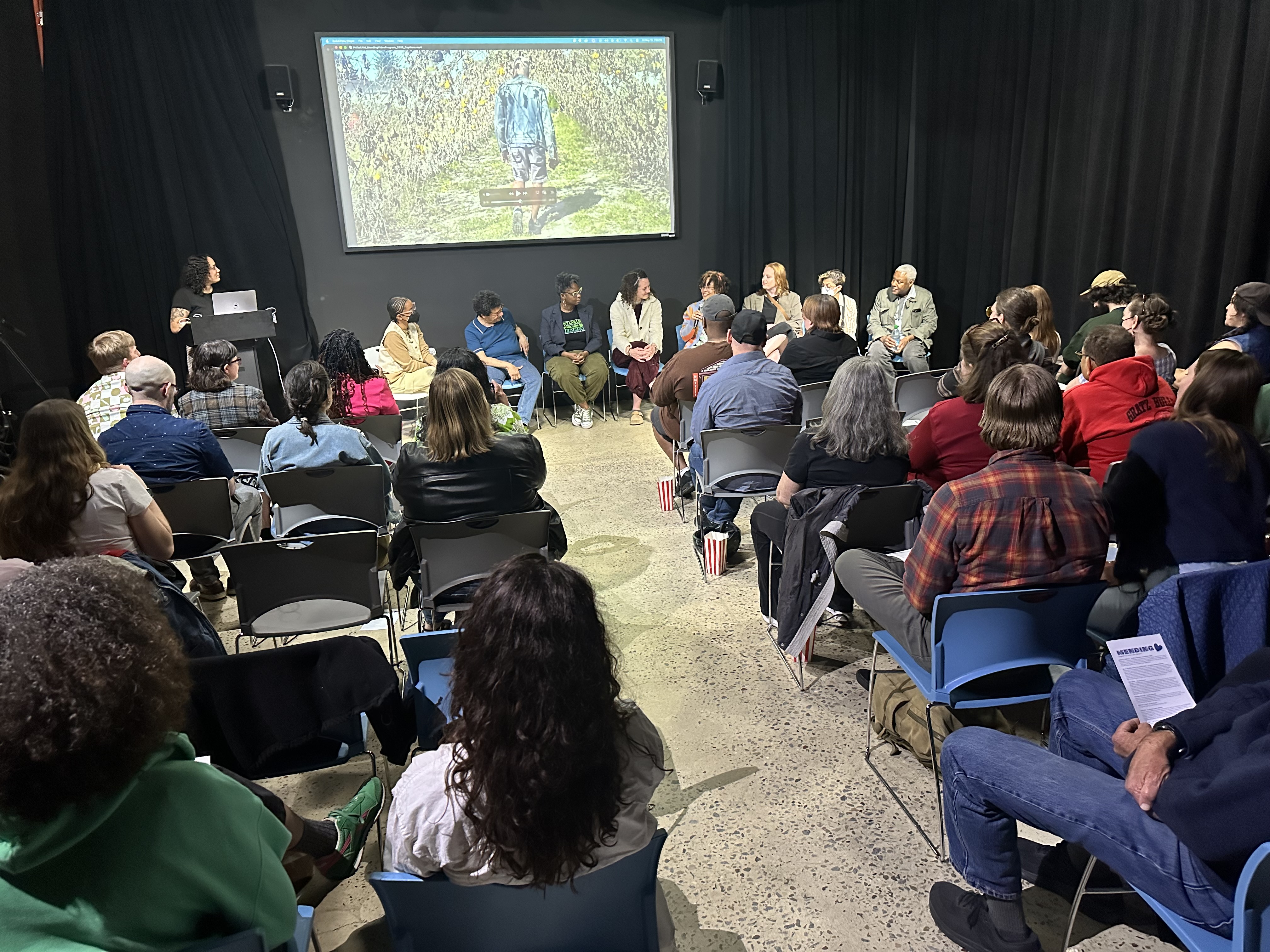A standing view of the Main TV studio where the screening was held. The point of view is from the back of the room with several rows of chairs facing the mounted screen and a row of chairs under the screening facing the audience. gabe is standing in the front of the room at the podium, all the chairs have people sitting with attention on the 8 filmmakers sitting in the front of the room.